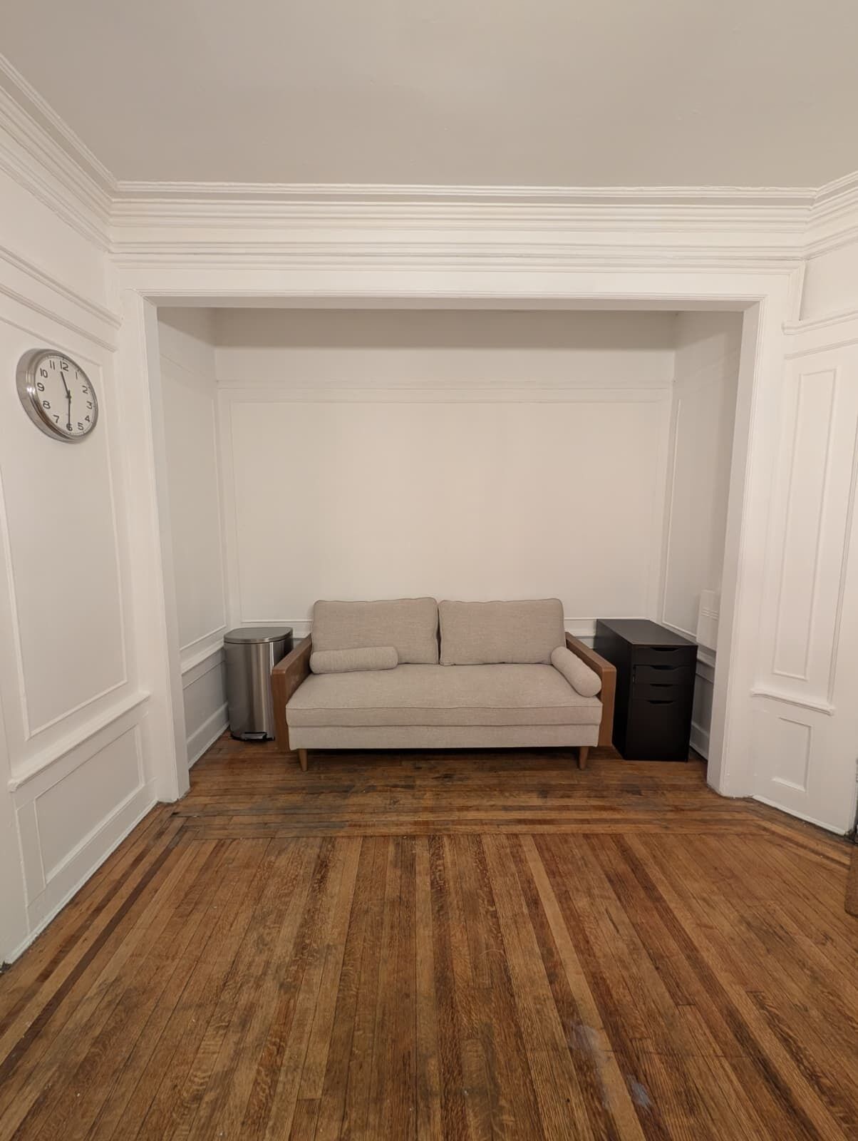 Une pièce avec un plancher en bois, un canapé beige, une poubelle et un classeur noir.