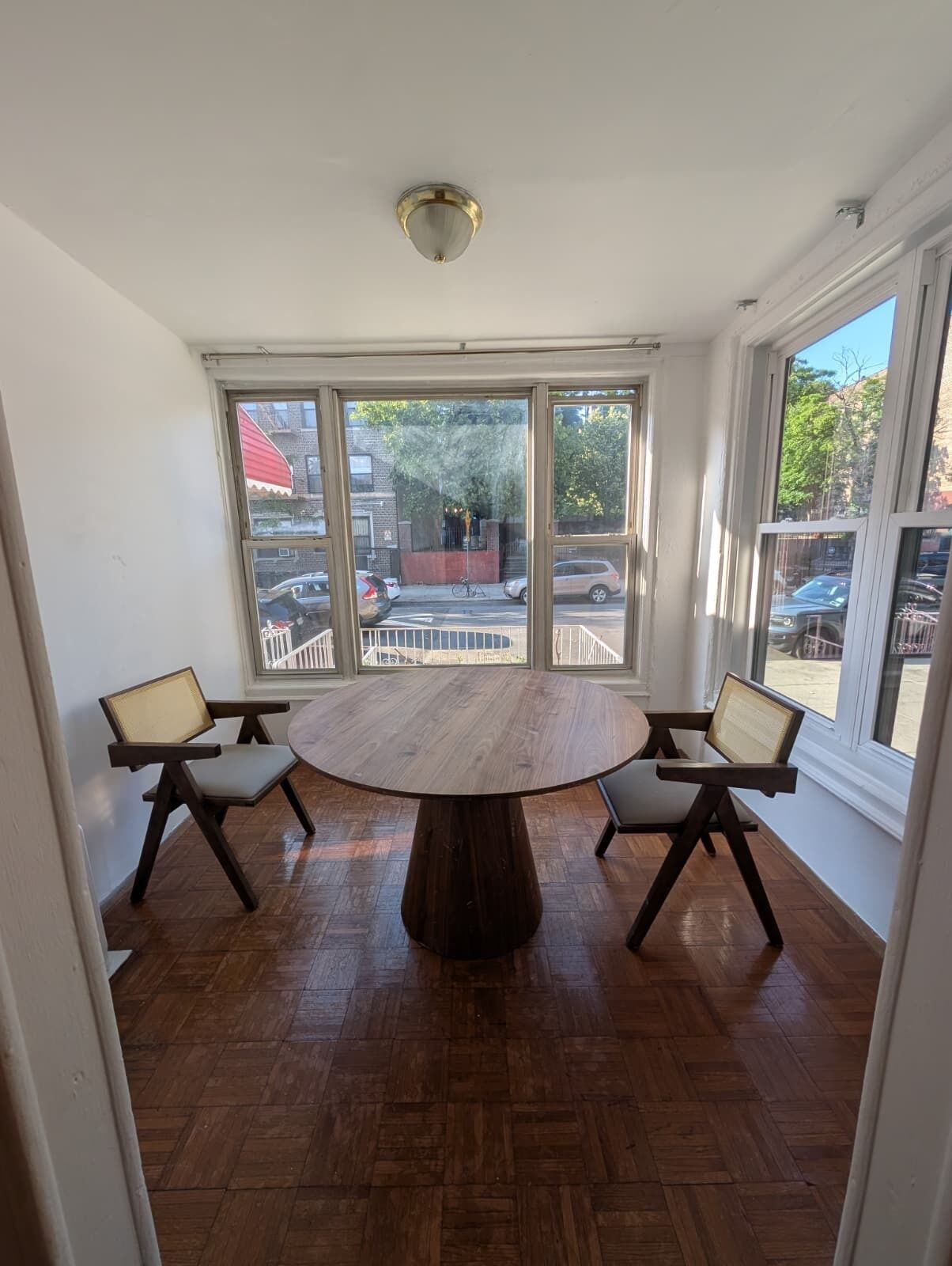 Une pièce avec une table ronde en bois, deux chaises, de grandes fenêtres et un luminaire au plafond.