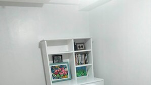 A white desk with a chair, a bookshelf with books and framed pictures, and a trash bin. - 2-BR w/ Full Kitchen & AC near Farmer’s Market, Araneta Coliseum & Greenhills (Quezon City)