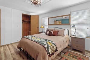 Ein Schlafzimmer mit Bett, Nachttisch, Spiegel, Kleiderschrank und einem Fenster mit Jalousien.