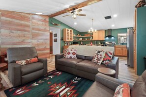 Un salón con sofá, sillón y alfombra. En el fondo, se ve una cocina con armarios de madera y un refrigerador.