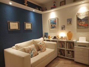 Ein Wohnzimmer mit blauer Wand, einem weißen Sofa mit gemusterten Kissen, einem Bücherregal und einem Rahmen mit der Aufschrift 'Willkommen'.
