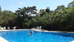En swimmingpool med fliset kant, omgivet af træer og hvide plaststole.