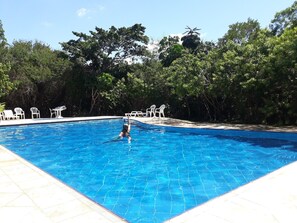 Ein Swimmingpool mit einer gefliesten Kante, umgeben von Bäumen und weißen Kunststoffstühlen.