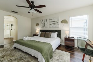 Un dormitorio con cama, ventilador de techo, mesita de noche, una silla y obras de arte en la pared.