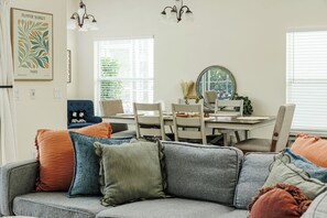 Sala de estar con un sofá gris, una mesa de comedor y sillas. Hay una ventana con persianas, una araña y un cuadro en la pared.