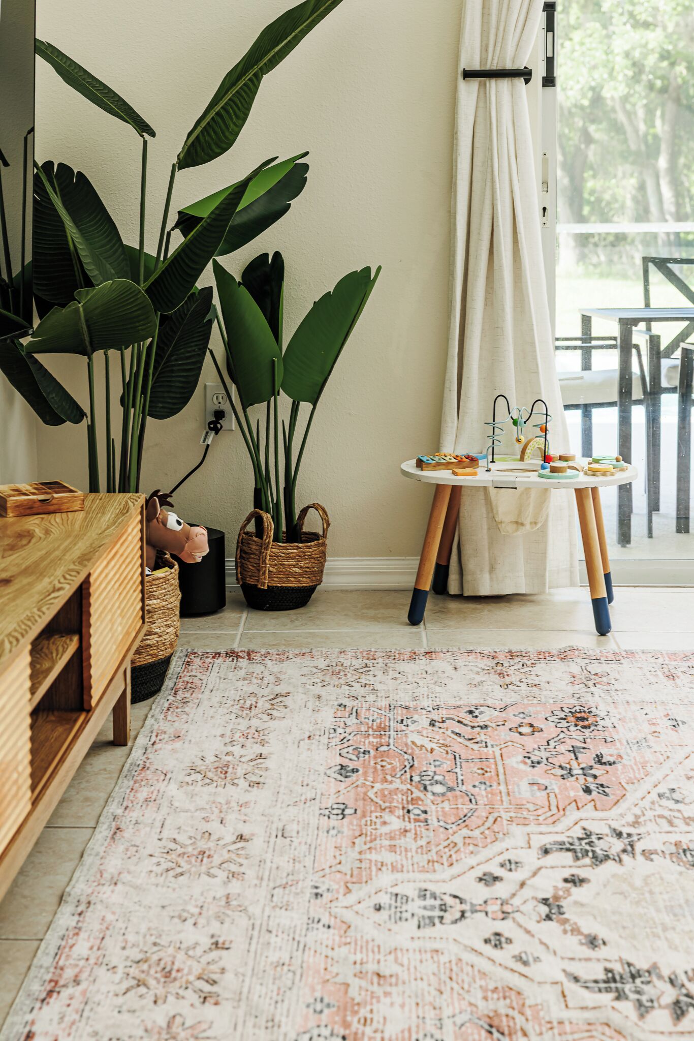 A room with a wooden cabinet, large green plants, a small table with a decorative item, and a patterned rug.