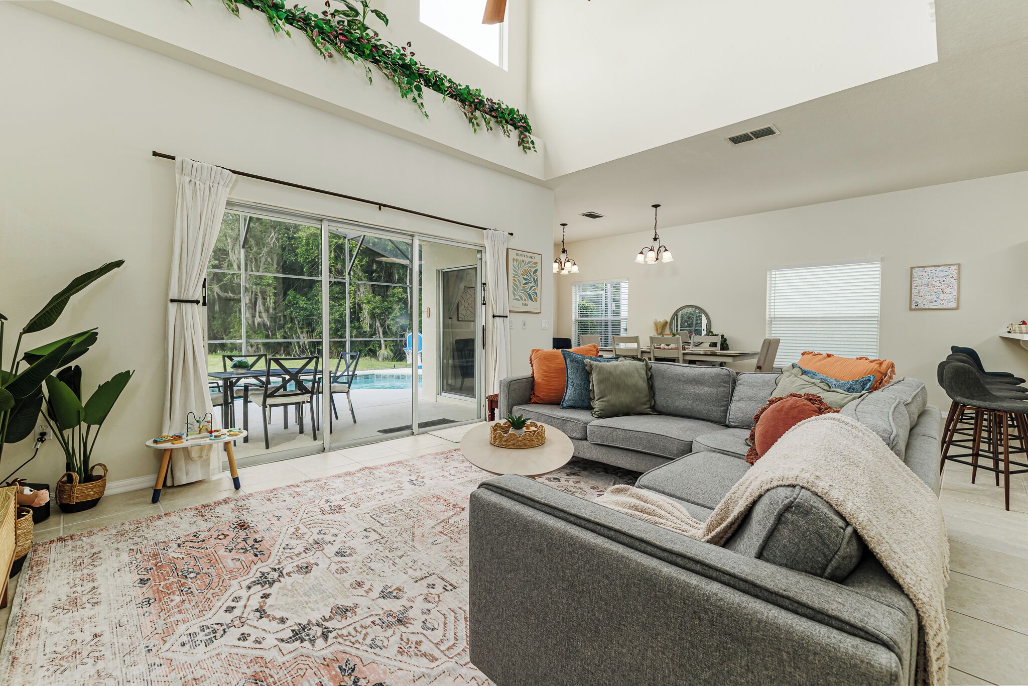 A spacious living area with a large sofa, a coffee table, and a patterned rug. There is a sliding glass door leading to an outdoor patio with a dining table and chairs.