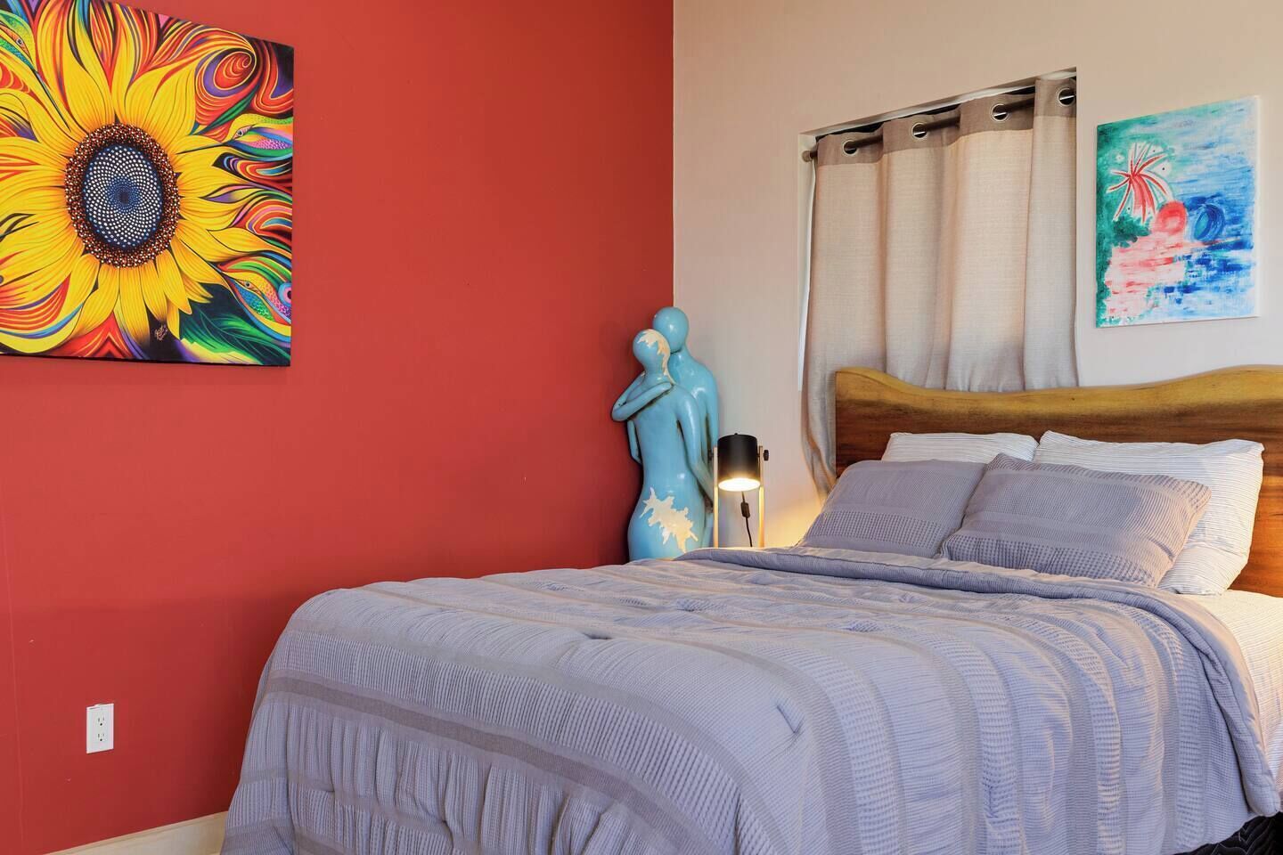 A bedroom with a red wall, a large bed with a wooden headboard, a blue statue, and two paintings.