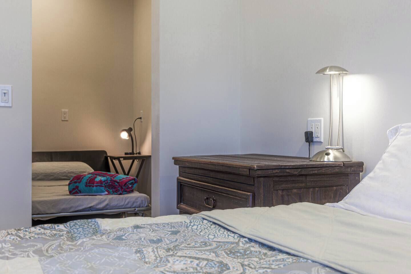 A bedroom with a bed, a bedside table with a lamp, a chair, and a desk with a lamp.