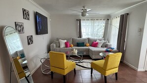 Una sala de estar con un sofá, dos sillas amarillas, un ventilador de techo y un televisor en la pared.