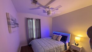Un dormitorio con una cama, una mesita de noche, un ventilador de techo y un respiradero en la pared.