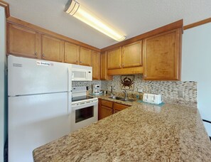 A kitchen with wooden cabinets, a refrigerator, a microwave, a toaster, and a coffee maker. - Whispering Sands II 204 - Second Row Complex (Oceanview unit) in Garden City Beach (Garden City Beach)