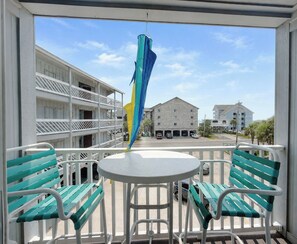 A balcony with a table and chairs, overlooking a street with buildings and parked cars. - Whispering Sands II 204 - Second Row Complex (Oceanview unit) in Garden City Beach (Garden City Beach)