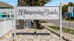 Un letrero de madera con el texto 'Whispering Sands' rodeado de piedras y palmeras.