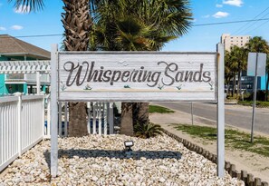 A wooden sign with the text 'Whispering Sands' surrounded by pebbles and palm trees. - Whispering Sands II 204 - Second Row Complex (Oceanview unit) in Garden City Beach (Garden City Beach)