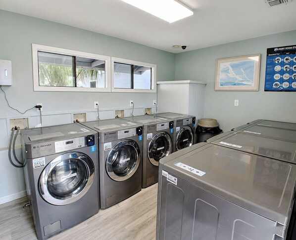 A laundry room with four front-loading washing machines, a large window, and a poster on the wall. - Whispering Sands II 204 - Second Row Complex (Oceanview unit) in Garden City Beach (Garden City Beach)