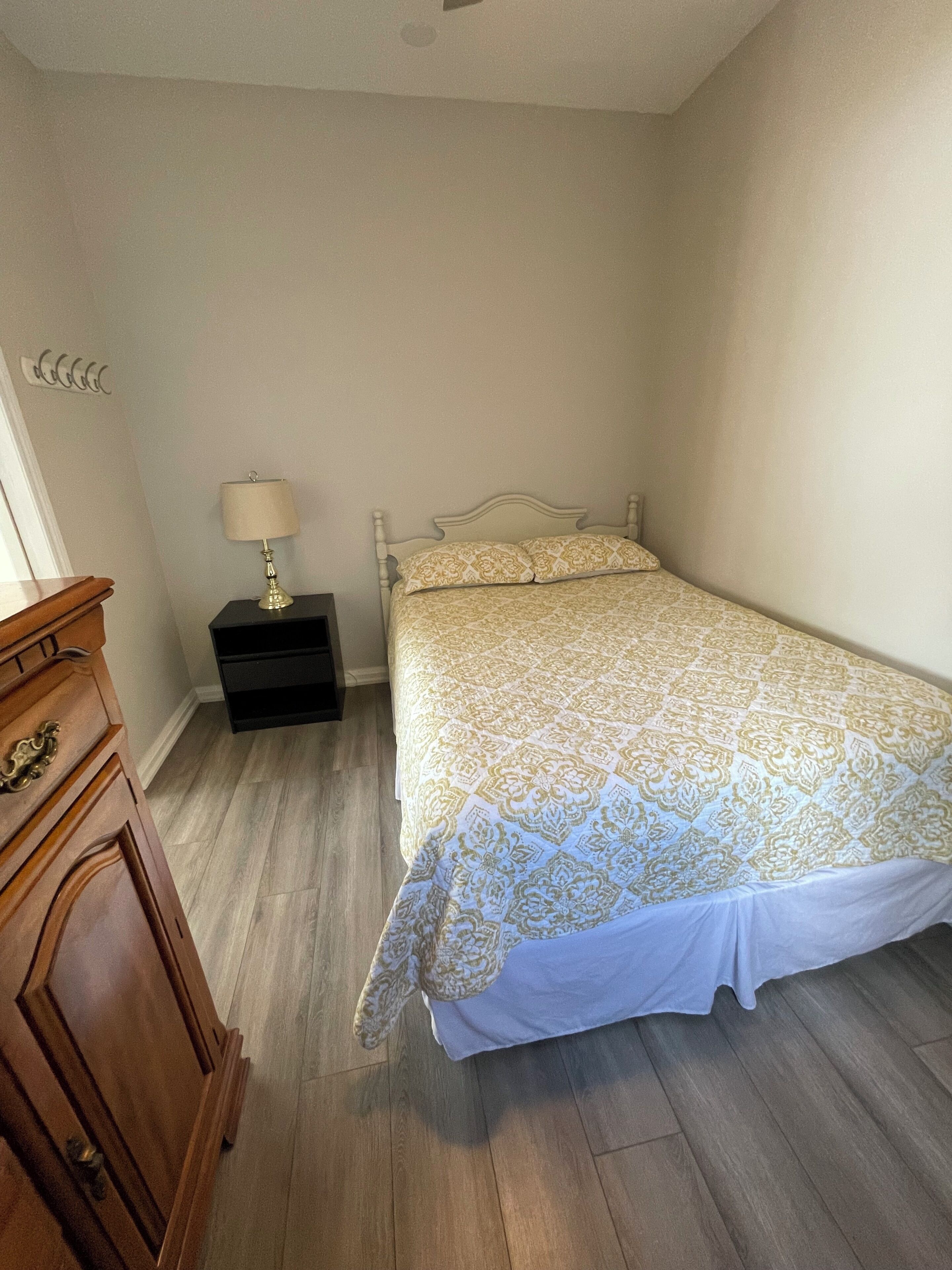 A bedroom with a bed, a nightstand with a lamp, a wooden dresser, and a light-colored floor.