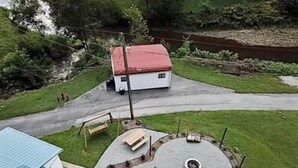 Ein Haus mit rotem Dach, einer gepflasterten Auffahrt, einer runden Terrasse mit Feuerstelle und einer Bank.