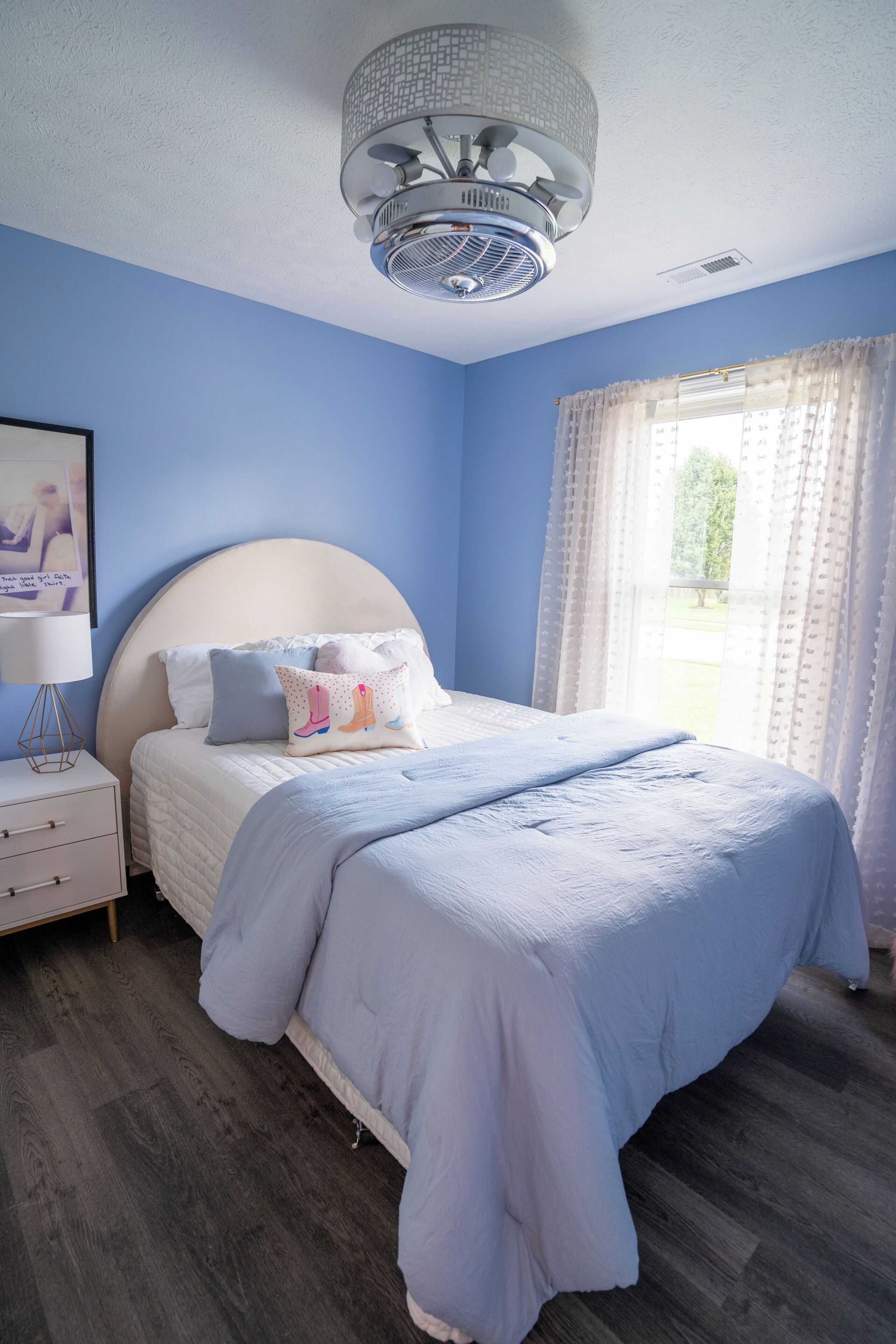 Un dormitorio con cama, mesita de noche, ventilador de techo y ventana con cortinas.