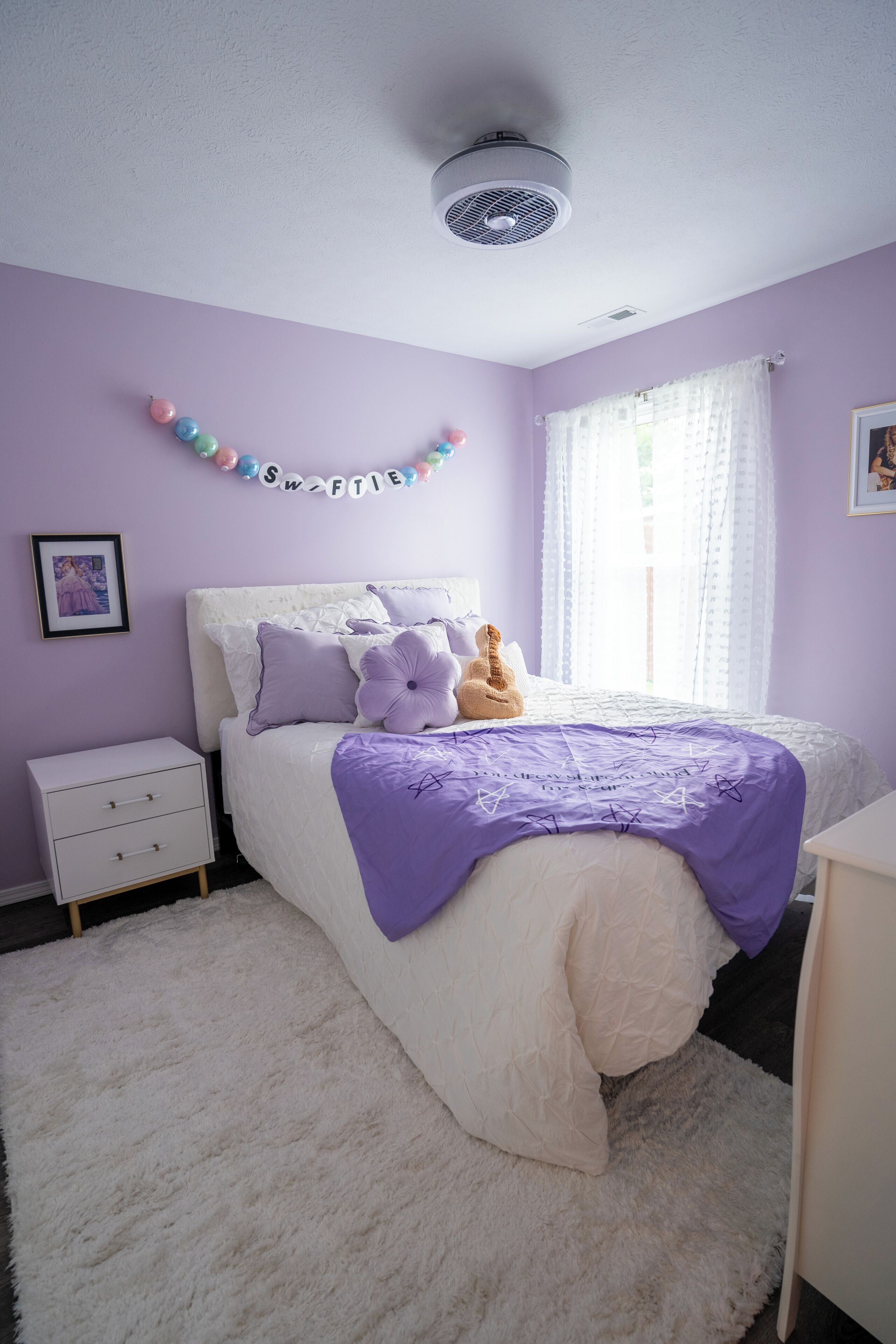A bedroom with a bed, a nightstand, a dresser, a window with curtains, and a wall-mounted air purifier.