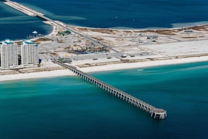 An aerial view of a coastal area with a long pier extending into the water, a beach, and several buildings including a hotel with multiple floors and a parking area. - Emerald Surf 204 (Navarre)