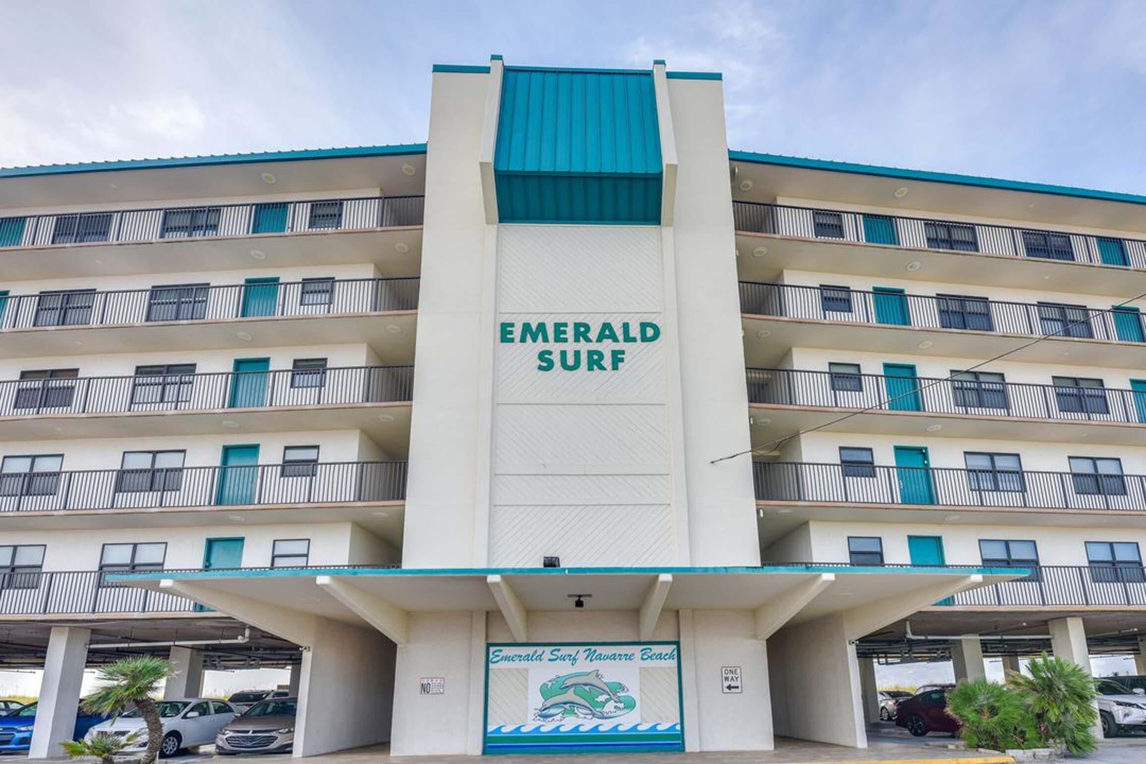A multi-story hotel with balconies, a prominent 'Emerald Surf' sign, and a parking area with cars.