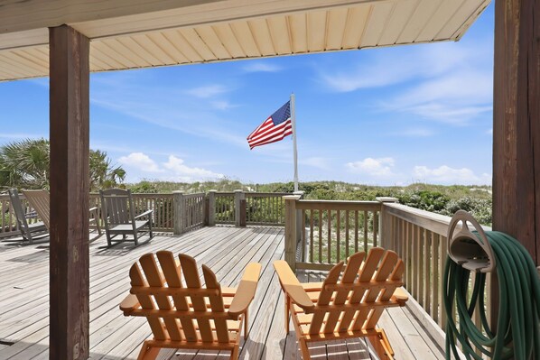 A wooden deck with two Adirondack chairs, a flag, and a view of the beach.