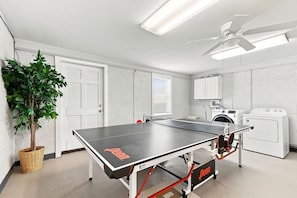 A room with a ping pong table, a ceiling fan, a washing machine, and a plant.