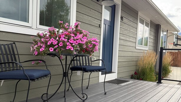 Una terraza de madera con dos sillas de metal y una mesa, una casa con puerta azul y flores rosas en una maceta.