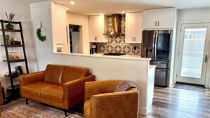 Una sala de estar moderna con un sofá y un sillón, una cocina con armarios blancos y un refrigerador de acero inoxidable, y un tapete estampado en el suelo.