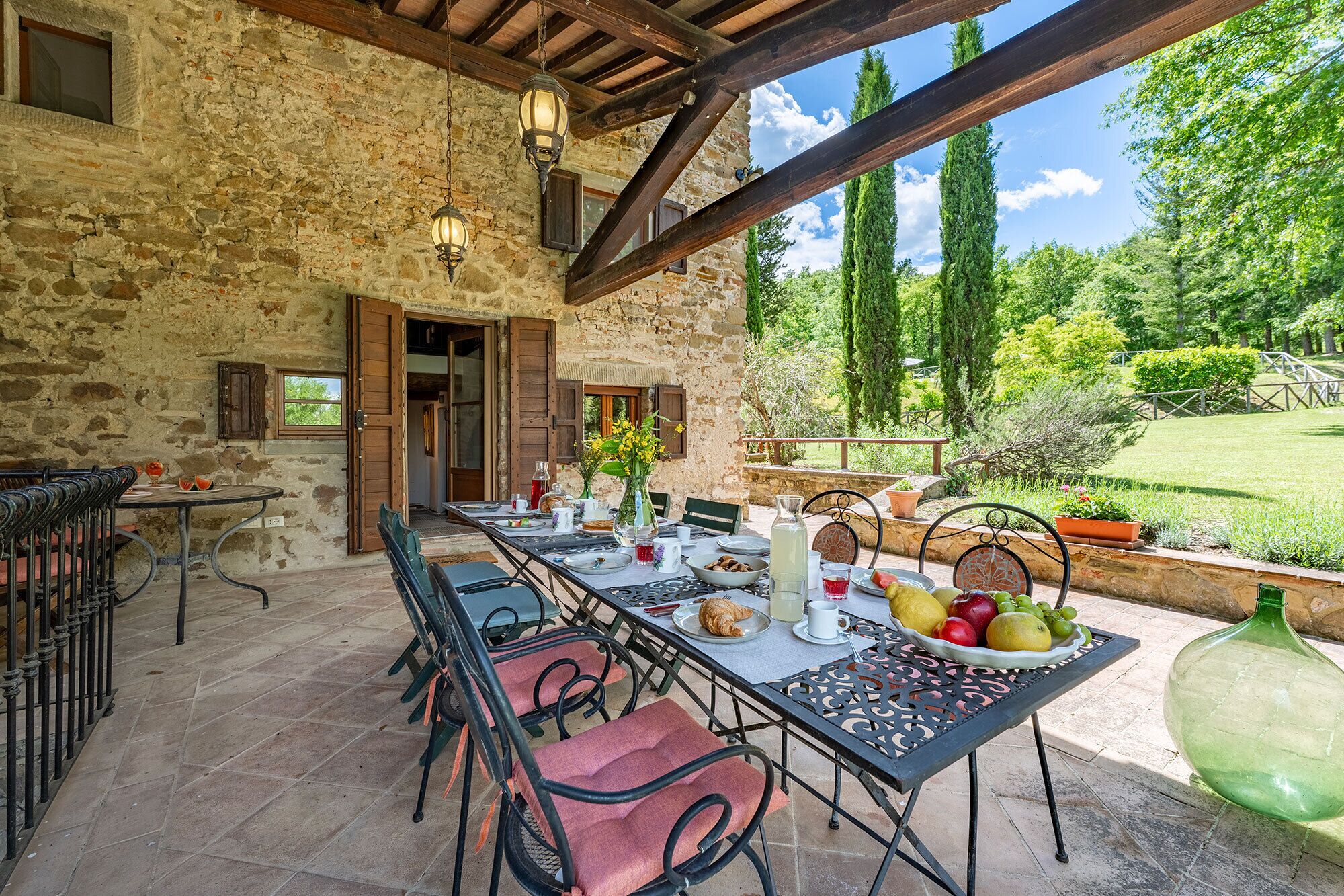 Un área de comedor al aire libre con paredes de piedra, vigas de madera y vista a un jardín exuberante.