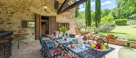 An outdoor dining area with stone walls, wooden beams, and a view of a lush garden.