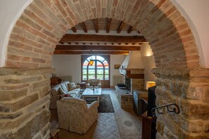 A cozy room with a stone archway, a fireplace, a sofa, and a table with a bottle and glasses. - Villa Palazzolo 8, Emma Villas (Anghiari)