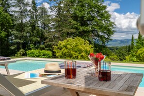 Una mesa de madera con bebidas y un sombrero de paja junto a una piscina con vistas panorámicas.