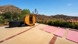 A sauna with a wooden exterior, surrounded by a patio with exercise mats, cacti, and a mountainous backdrop. - Desert Sunsets Wellness Villa - Pool, Sauna, Spa, Pool Table, Firepit, Yoga, Steam Room & Stunning Views (Yucca Valley)