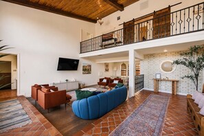 A spacious living area with a high ceiling, a balcony with a wooden railing, a flat-screen TV mounted on the wall, and a patterned rug on the floor. - Desert Sunsets Wellness Villa · Pool, Sauna, Views (Yucca Valley)