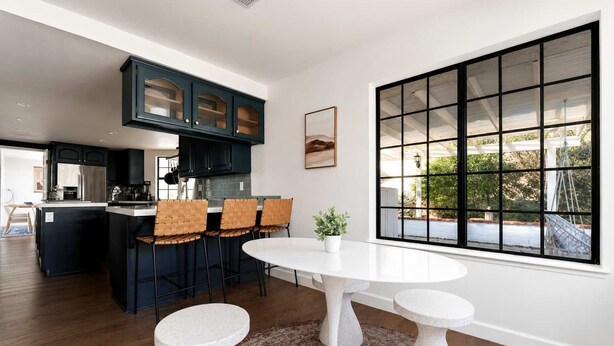 A modern kitchen with dark cabinets, a central island, and a dining area with a white table and chairs. - Desert Sunsets Wellness Villa - Pool, Sauna, Spa, Pool Table, Firepit, Yoga, Steam Room & Stunning Views (Yucca Valley)