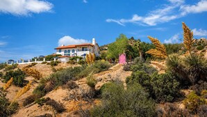 A large house with multiple balconies, surrounded by a variety of desert vegetation and a clear blue sky. - Desert Sunsets Wellness Villa - Pool, Sauna, Spa, Pool Table, Firepit, Yoga, Steam Room & Stunning Views (Yucca Valley)