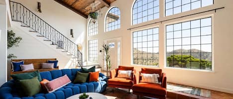 A spacious living room with a blue sofa, orange armchairs, and a round coffee table. A staircase with a railing is visible in the background.