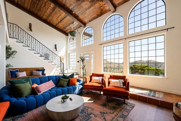 A spacious living room with a blue sofa, orange armchairs, and a round coffee table. A staircase with a railing is visible in the background.