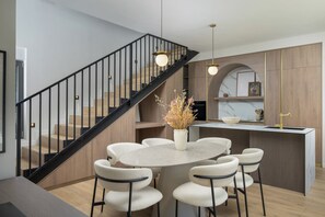 Eine moderne Küche mit einer zentralen Kochinsel, einem Essbereich mit sechs Stühlen, einer Treppe mit schwarzen Geländern und Pendelleuchten.