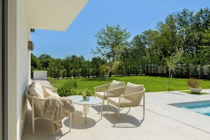 Eine Terrasse im Freien mit weißen Korbmöbeln, einem runden Tisch und Blick auf ein Schwimmbad und Grünflächen.