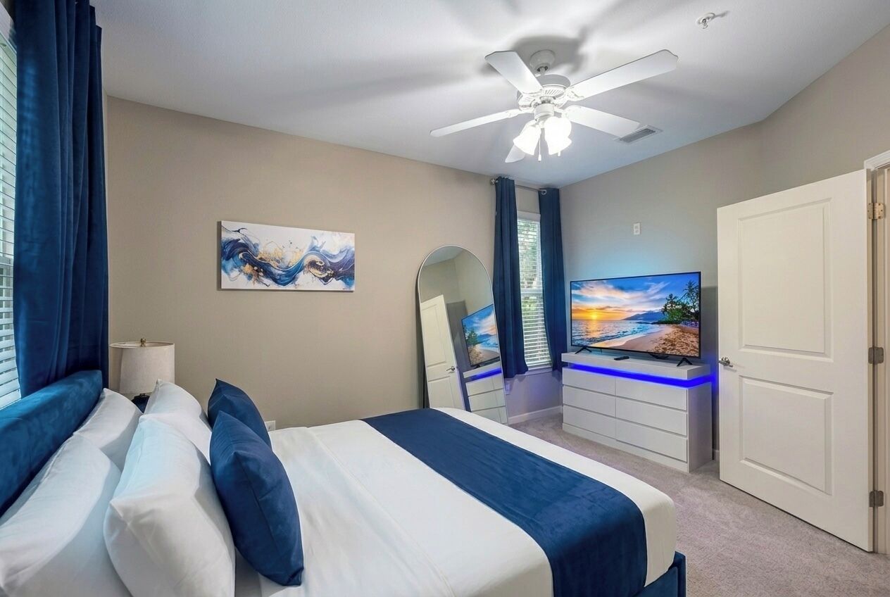 Ein Schlafzimmer mit einem Bett, einem Deckenventilator, einem Fernseher, einem Schminktisch und einer Tür.