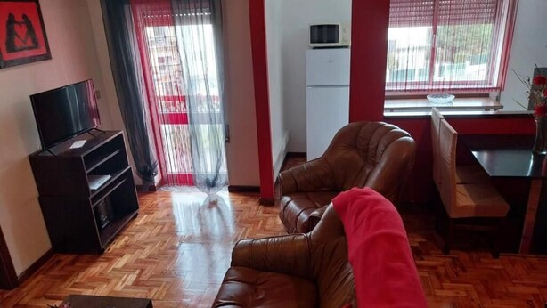 Un salón con un sofá de cuero marrón, un televisor sobre un mueble bajo, suelo de madera en espiga, un refrigerador blanco y una ventana con cortinas.