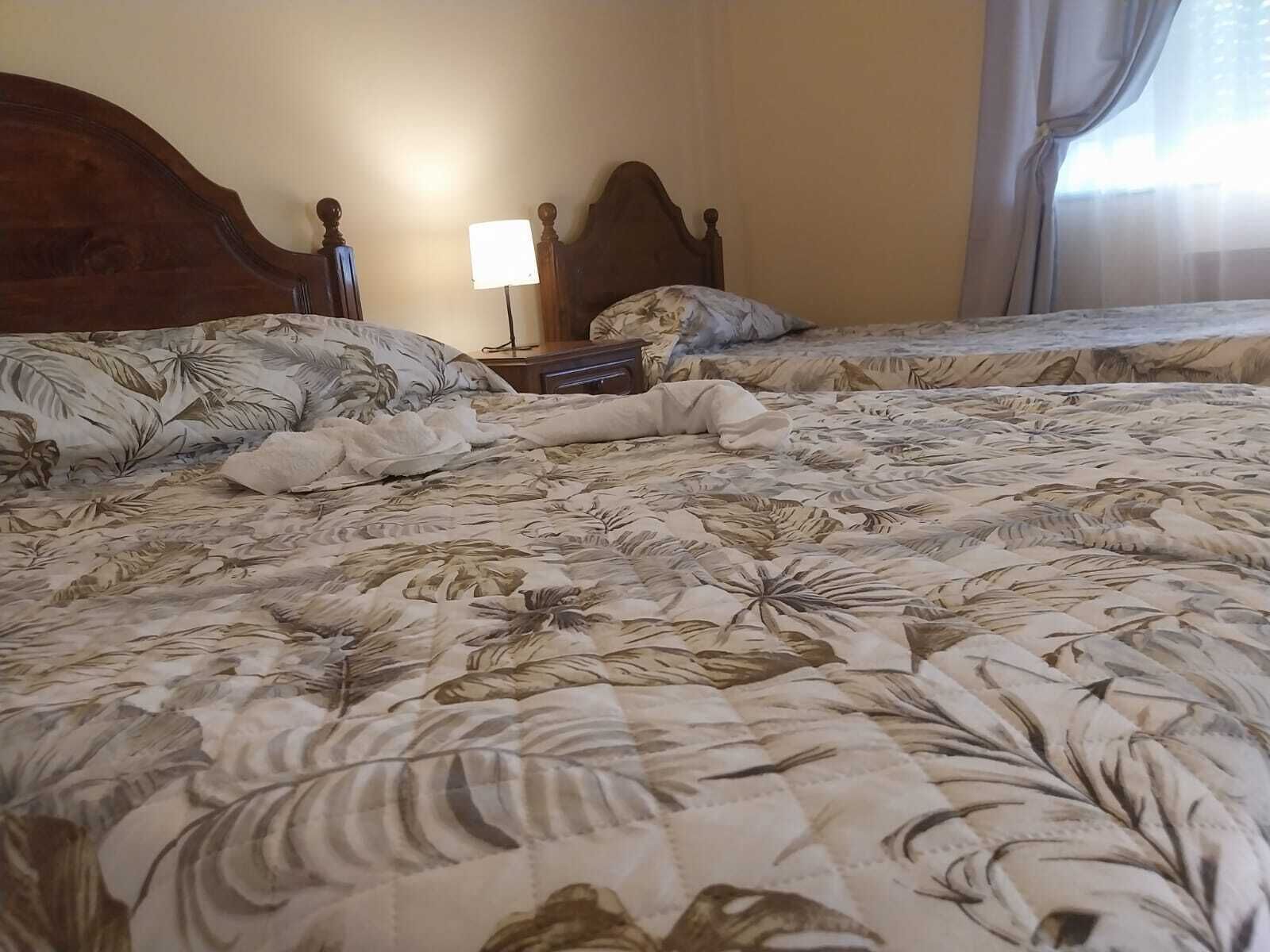 Quarto de hotel com duas camas, uma mesa de cabeceira, uma luminária e uma janela com cortinas.