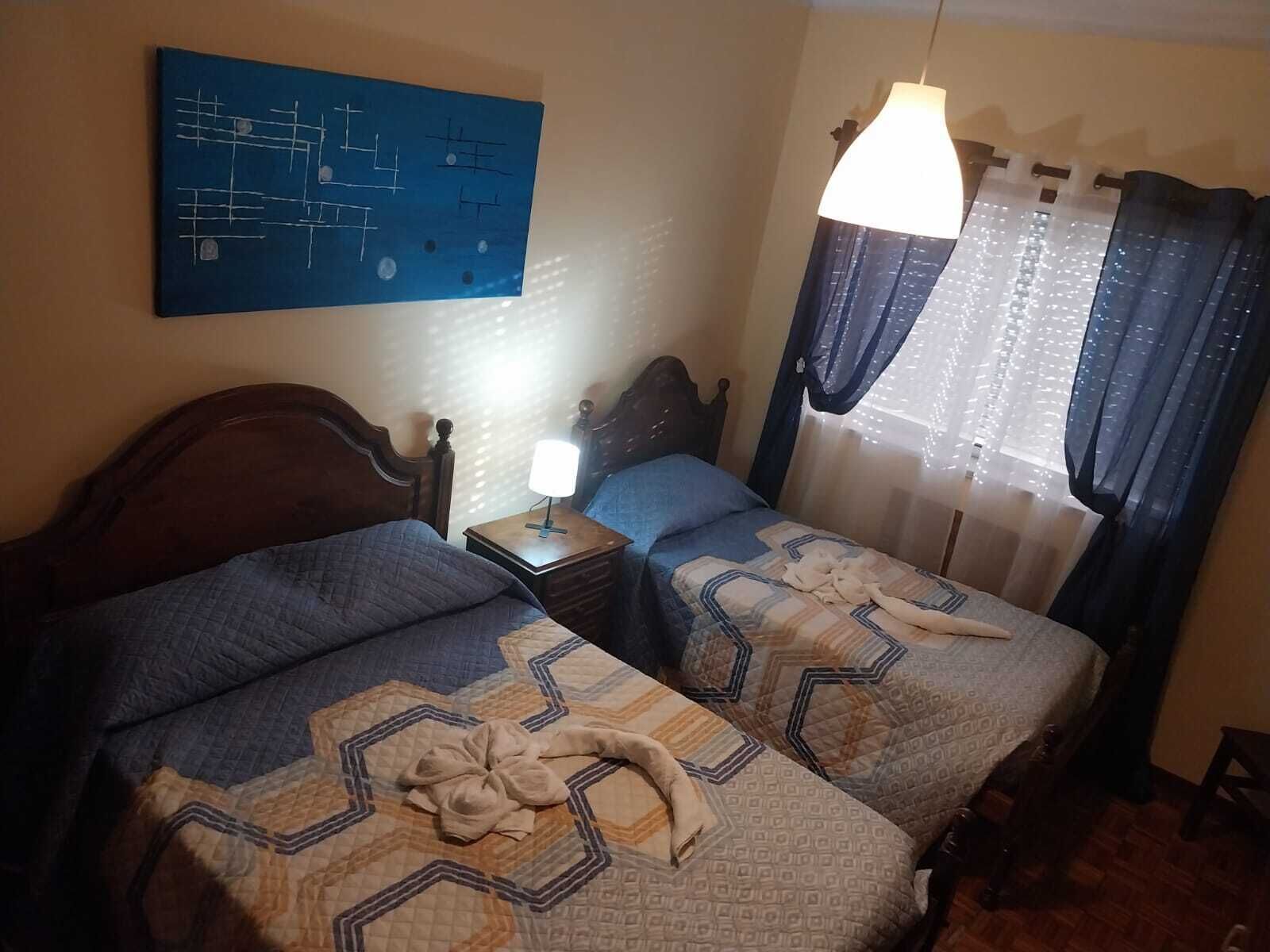 A hotel room with two beds, a nightstand with a lamp, a blue abstract painting on the wall, and a window with curtains.