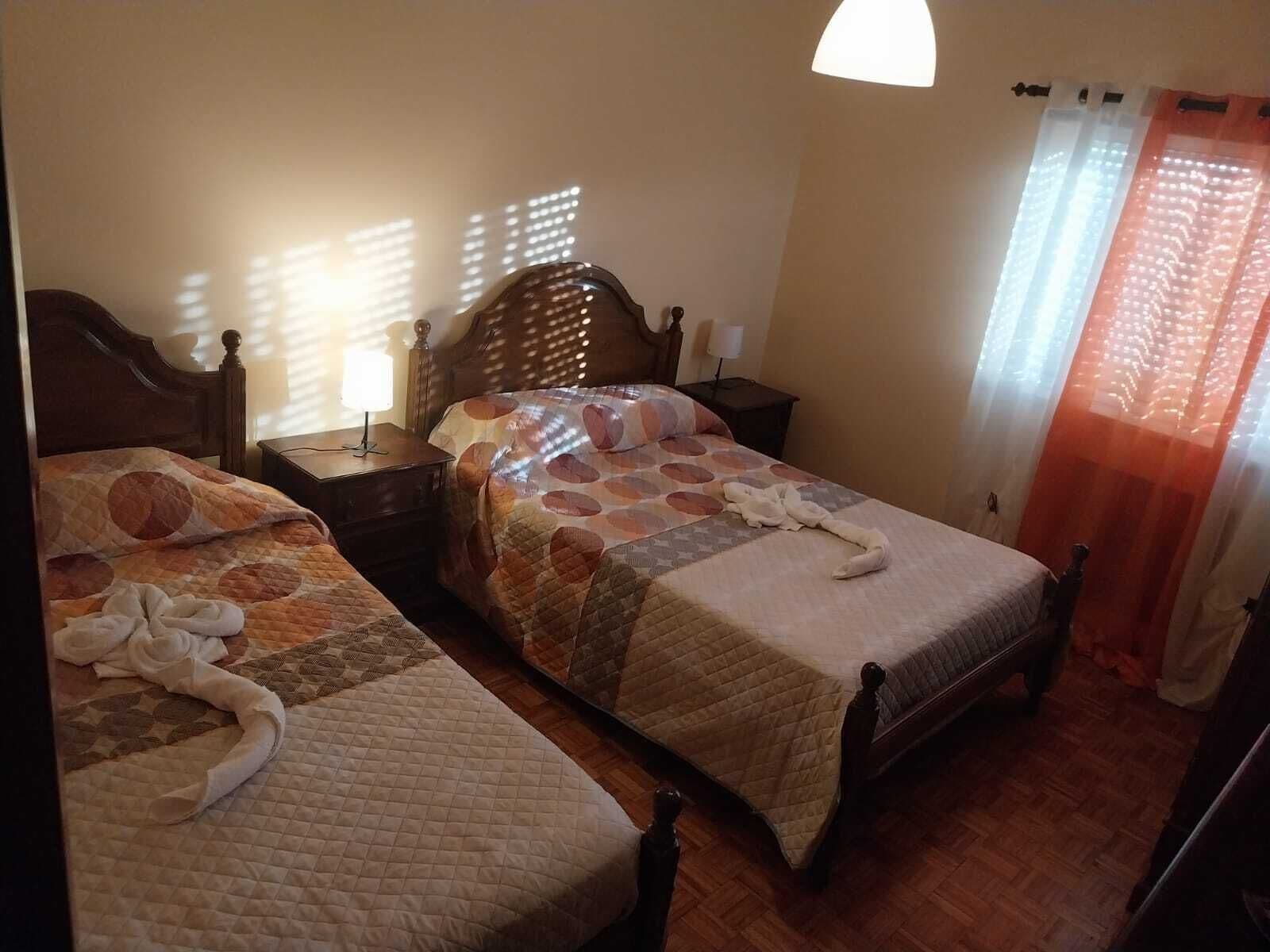 Een hotelkamer met twee bedden, houten hoofdeinden, een nachtkastje met een lamp en een raam met gordijnen.