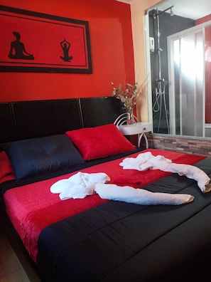 Una cama con ropa de cama rojo y negro, un florero blanco con flores y una zona de ducha visible a través de una puerta de cristal.