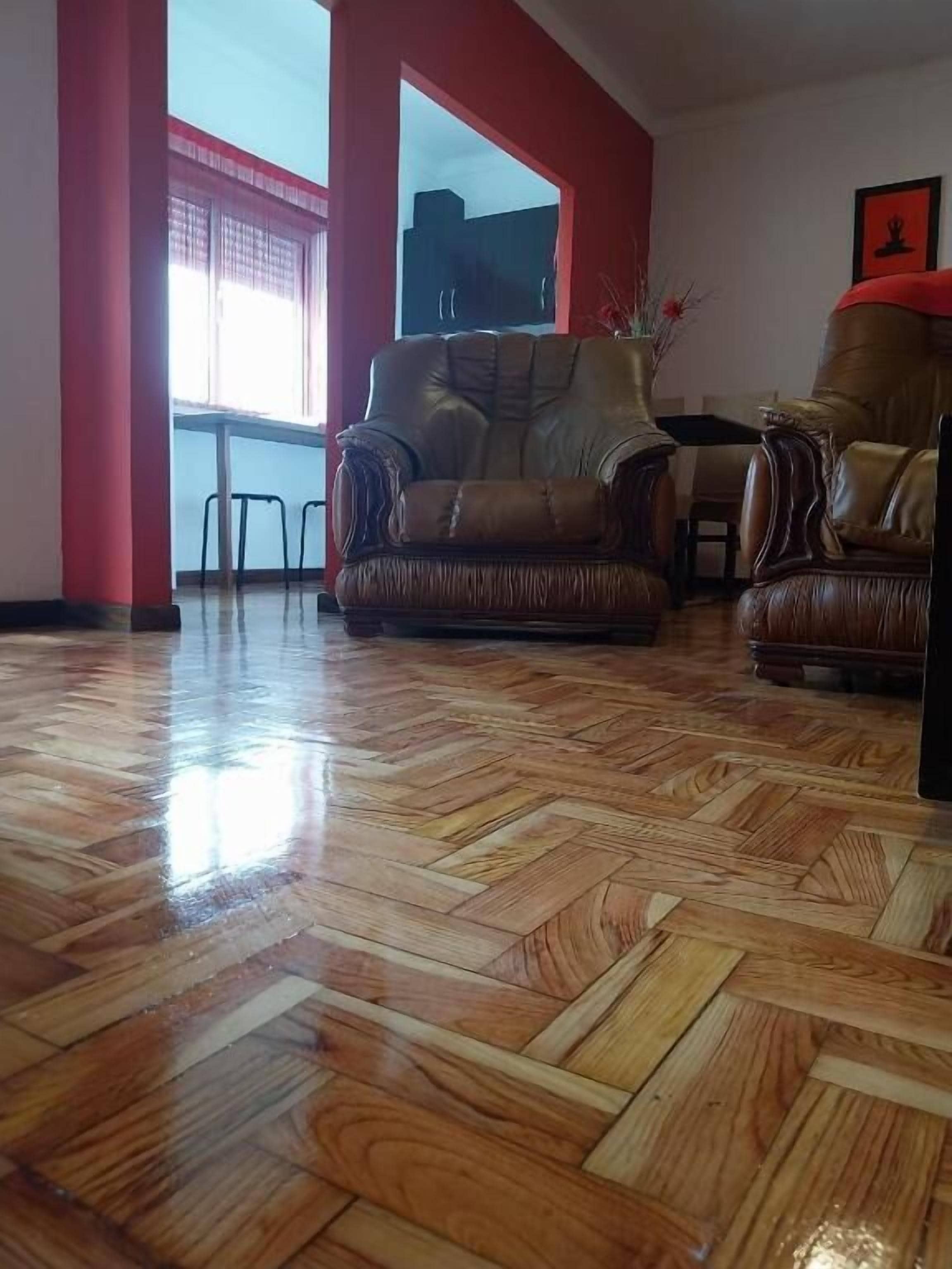 A room with a polished wooden floor, two leather sofas, a dining table, and a large window with red framing.